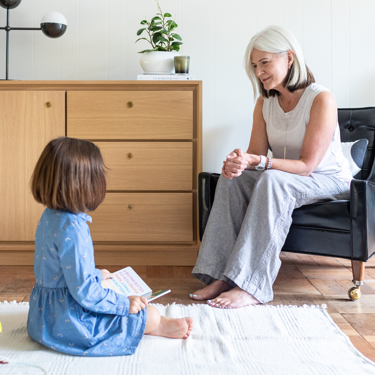 a woman with her young niece is sitting next to the Forti Goods Eleanor smart locking cabinet in Oak and clear finish with brass hardware pulls.