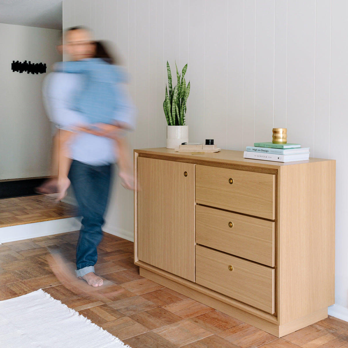 dad is carrying his young daughter across a living room, past the Forti Goods' Eleanor smart locking cabinet in oak with a clear finish.